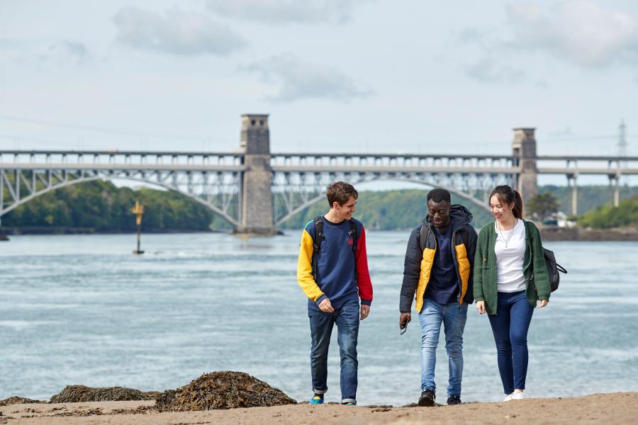 Three students walking along the beach with the Britannia bridge in the background